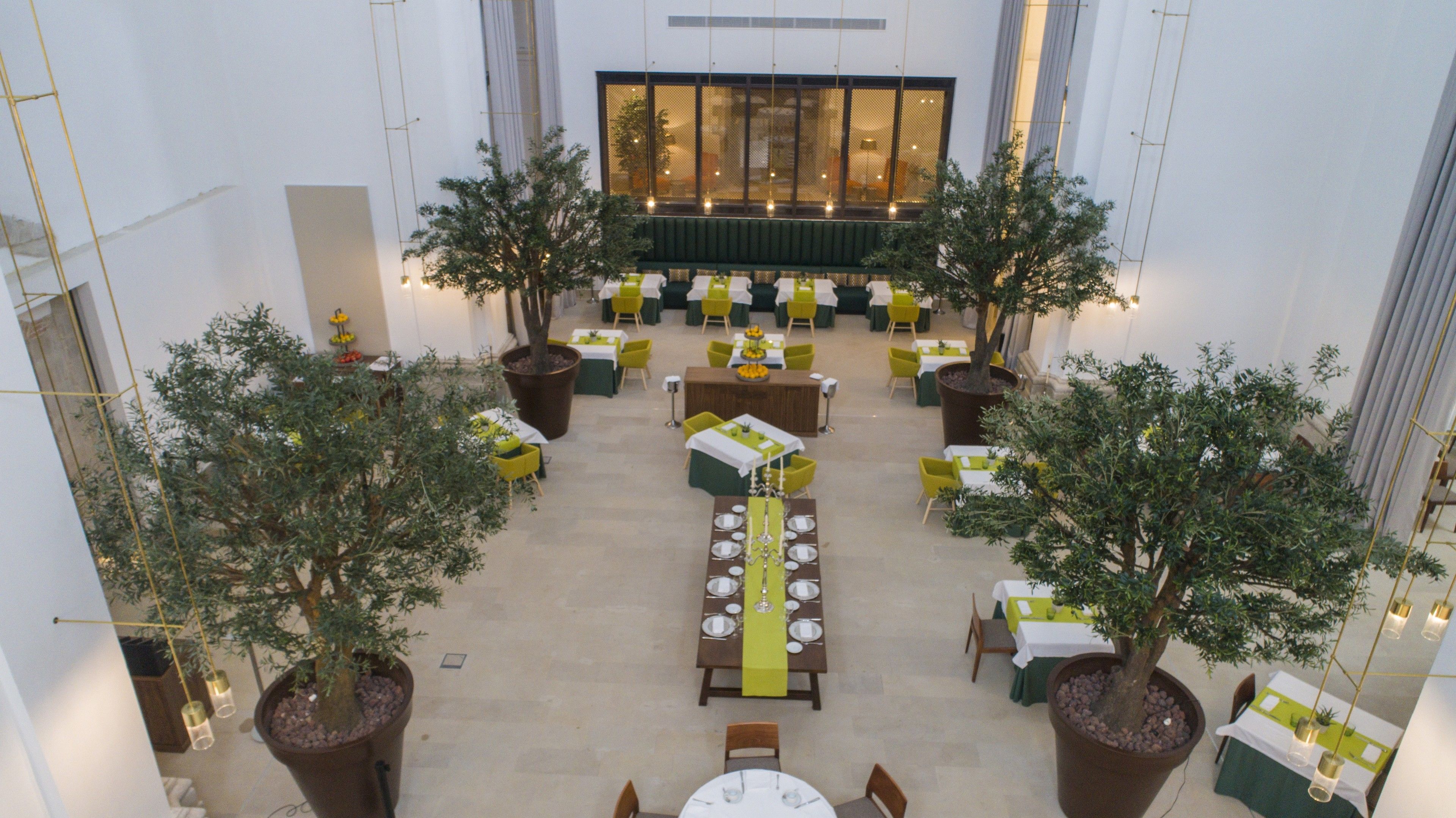 A spacious indoor dining area with multiple tables and chairs, surrounded by large potted trees.