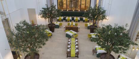 A spacious indoor dining area with multiple tables and chairs, surrounded by large potted trees.