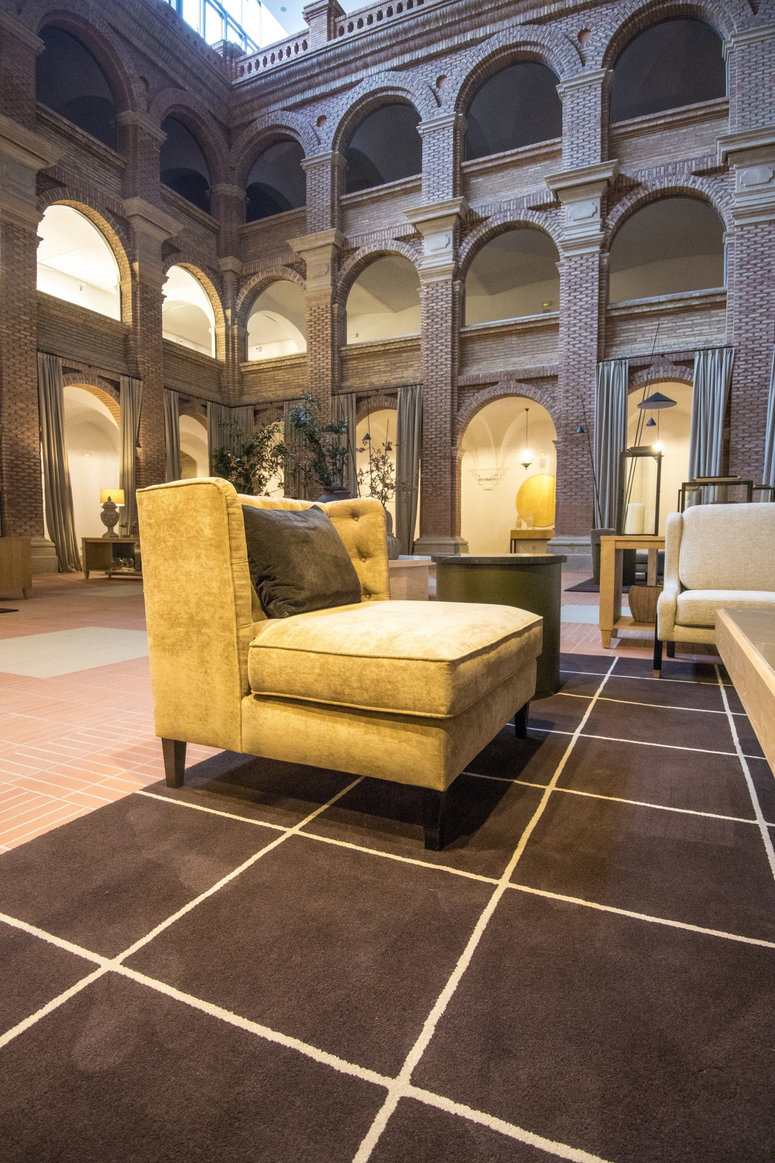 Un lobby amplio con techo alto, que presenta un sofá grande y mullido junto a una mesa de centro. El lobby tiene una pared de ladrillo con ventanas arqueadas y suelo de baldosas.