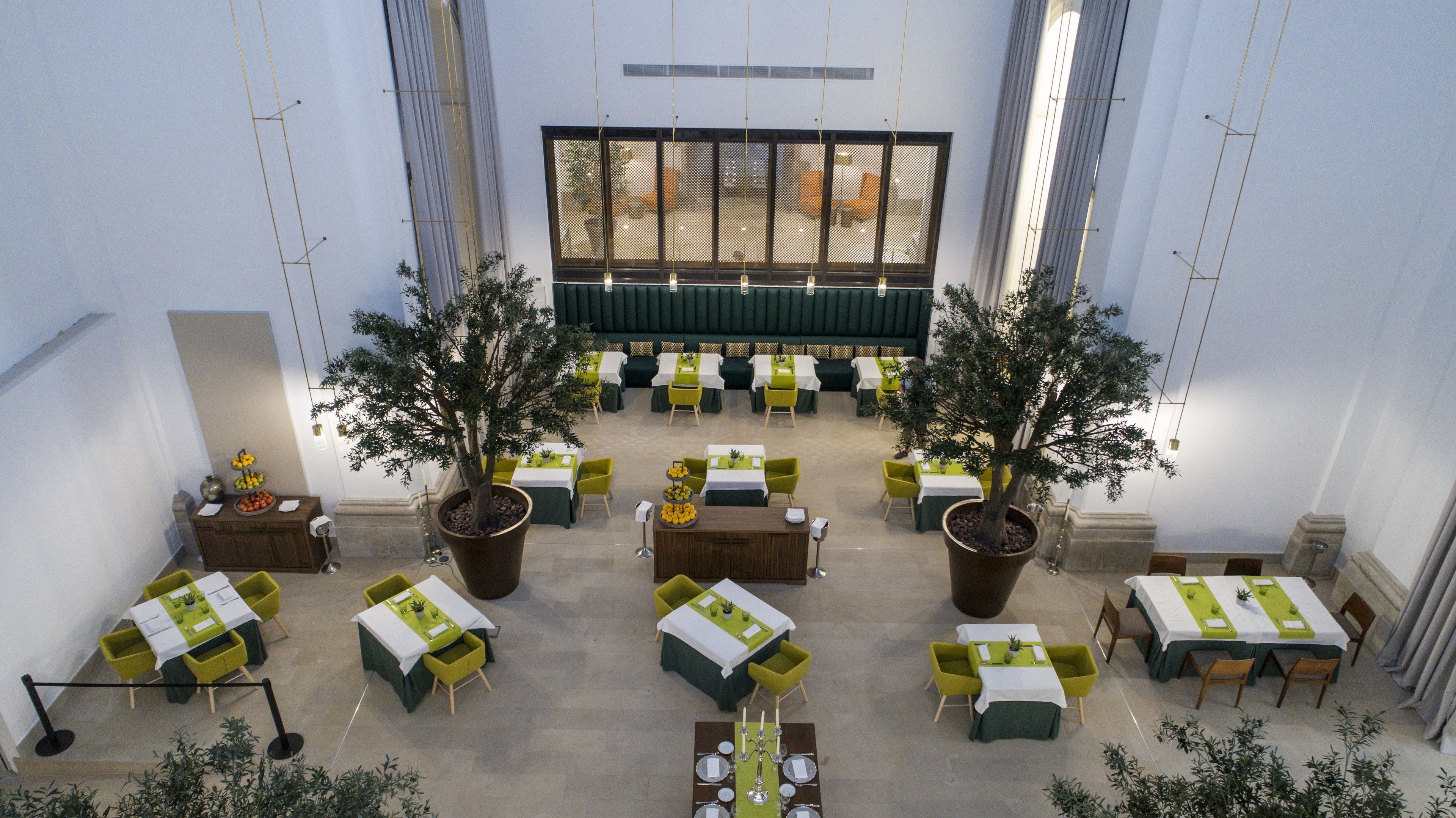A spacious indoor dining area with multiple tables and chairs, large potted plants, and a high ceiling with hanging lights.