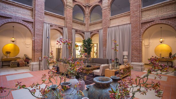 A spacious lobby with a high ceiling, brick walls, and large windows. There are several seating areas with sofas and tables, and decorative vases with flowers. - Parador de Lleida (Lleida)