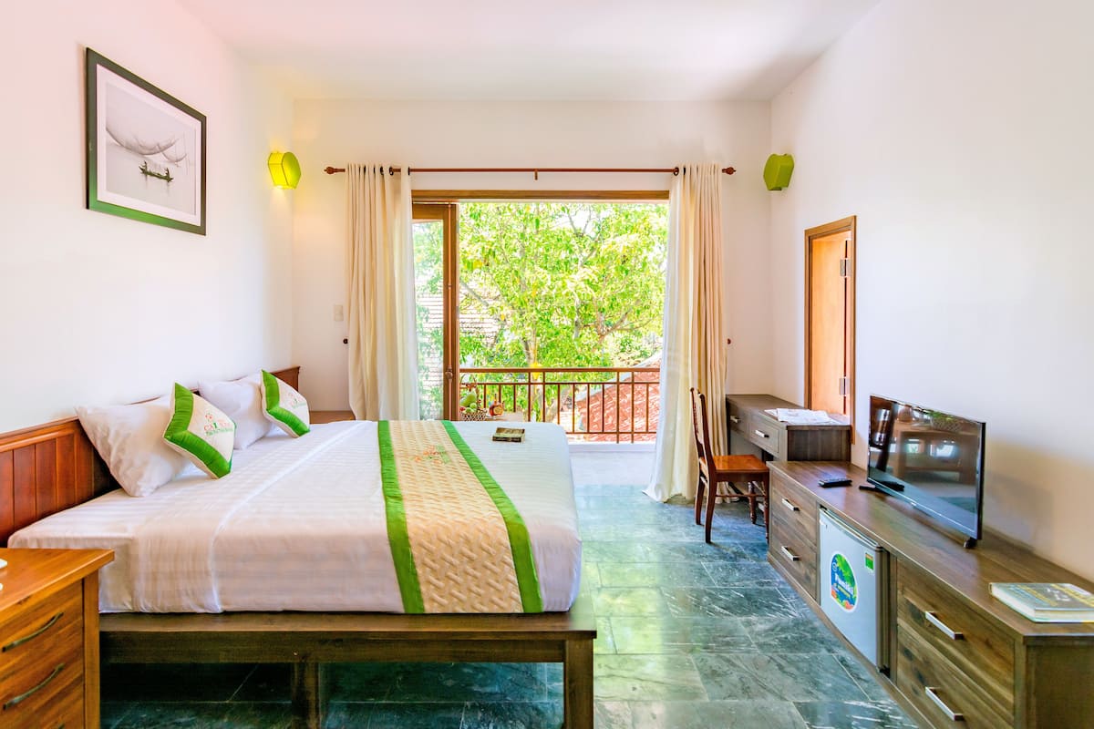 A hotel room with a bed, a desk, a television, and a balcony with a view of trees.