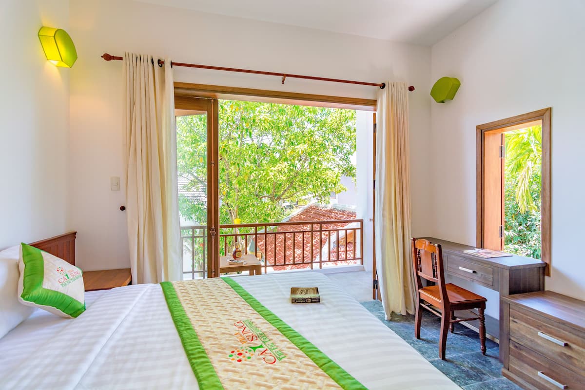 A hotel room with a bed, a desk, and a chair. There is a balcony with a view of trees and a red-roofed building.
