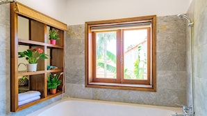 A bathroom with a large bathtub, a window with a view of greenery, and shelves with plants and towels. - Fig Tree Boutique Villa (Da Nang)