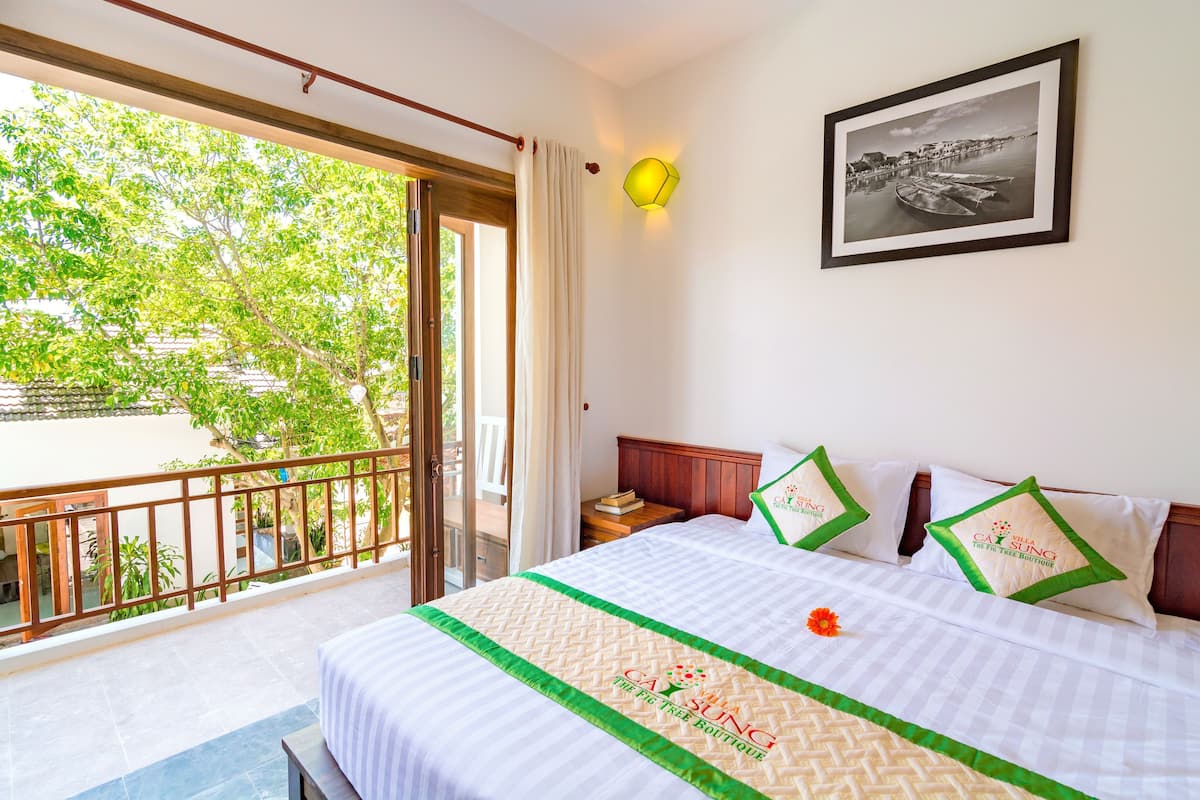 A hotel room with a bed, a framed photo on the wall, and a balcony with a view of greenery.