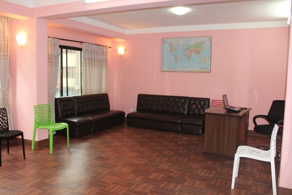 Une pièce avec un parquet en bois disposé en motif de chevron, un canapé en cuir noir, un fauteuil vert, un fauteuil blanc, un bureau en bois avec un ordinateur portable, et une carte du monde accrochée au mur.
