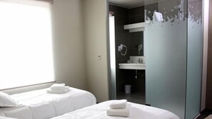 Une chambre d’hôtel avec deux lits, une salle de bain dotée d’une douche à parois de verre, et donnant sur la chambre voisine.