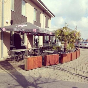 An outdoor cafe with tables and chairs, a large umbrella, and a building in the background. - Hotel Otus (Wetteren)