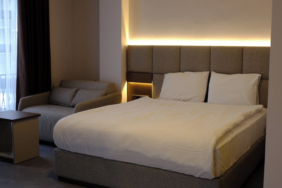 A modern hotel room with a bed, a sofa, a nightstand, and a wall-mounted light.