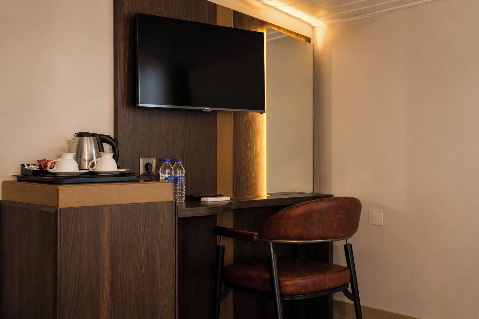 Habitación de hotel con un escritorio de madera, un televisor de pantalla plana montado encima, un sillón de cuero marrón y una minibar con cafetera, botellas de agua y un control remoto.