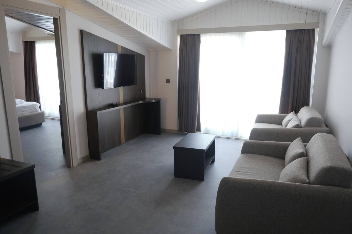 A modern living room with a sofa set, a coffee table, a TV mounted on the wall, and a sliding door leading to a bedroom.
