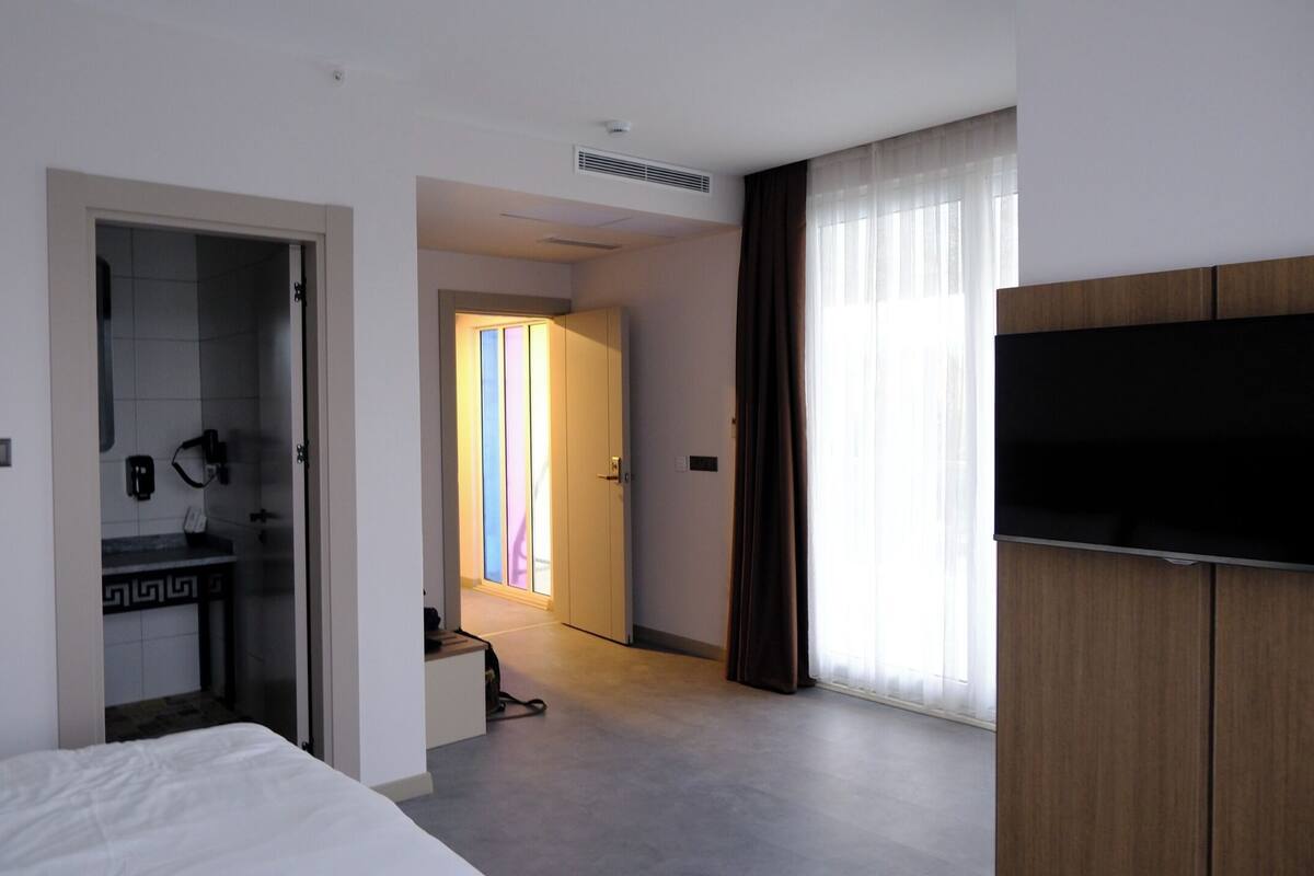 A hotel room with a bed, a TV, a bathroom with a mirror and towel rack, and a hallway leading to another room.