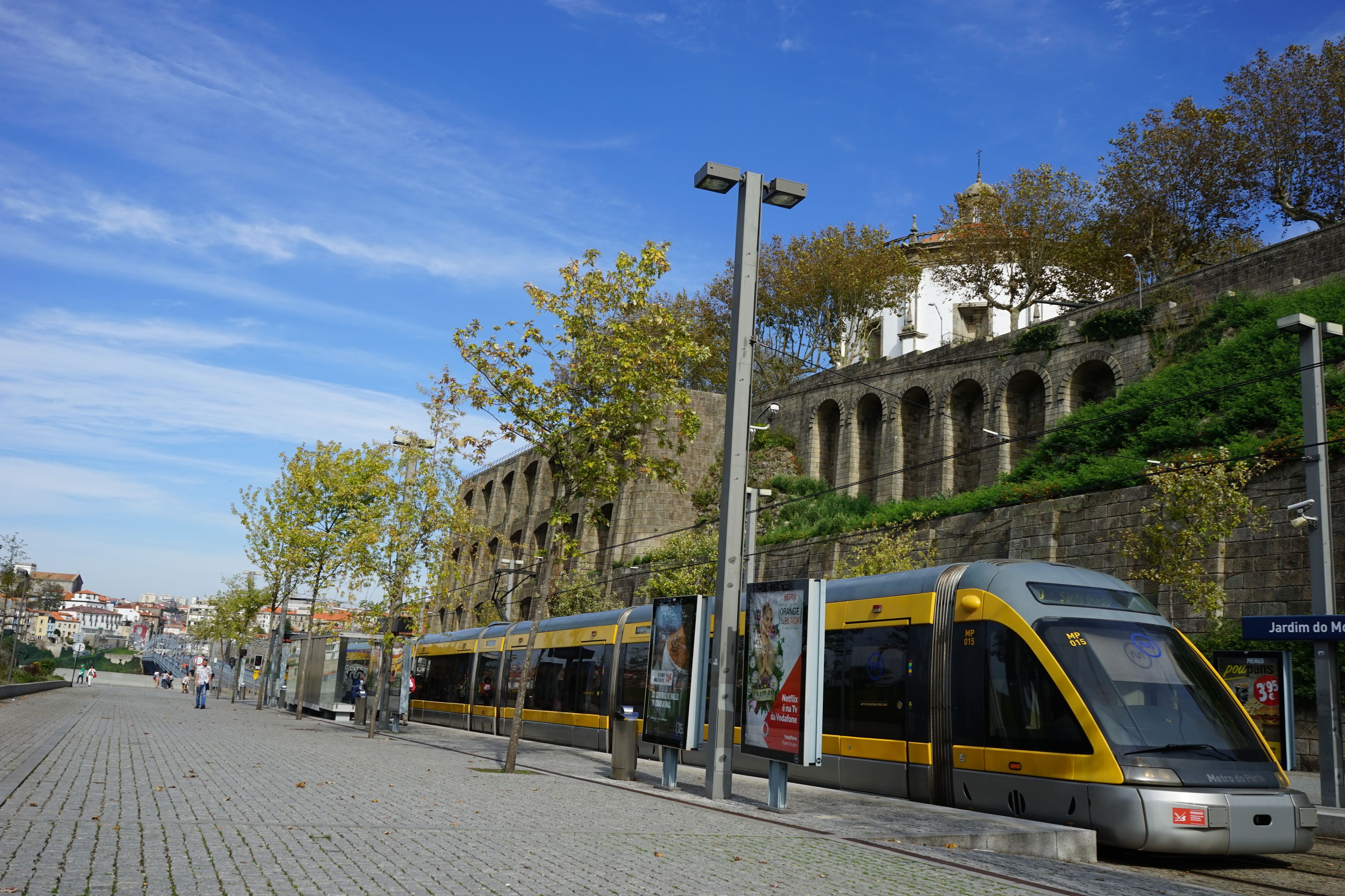 A modern train station with a yellow and gray train, a paved platform, and a historic building with arches in the background.