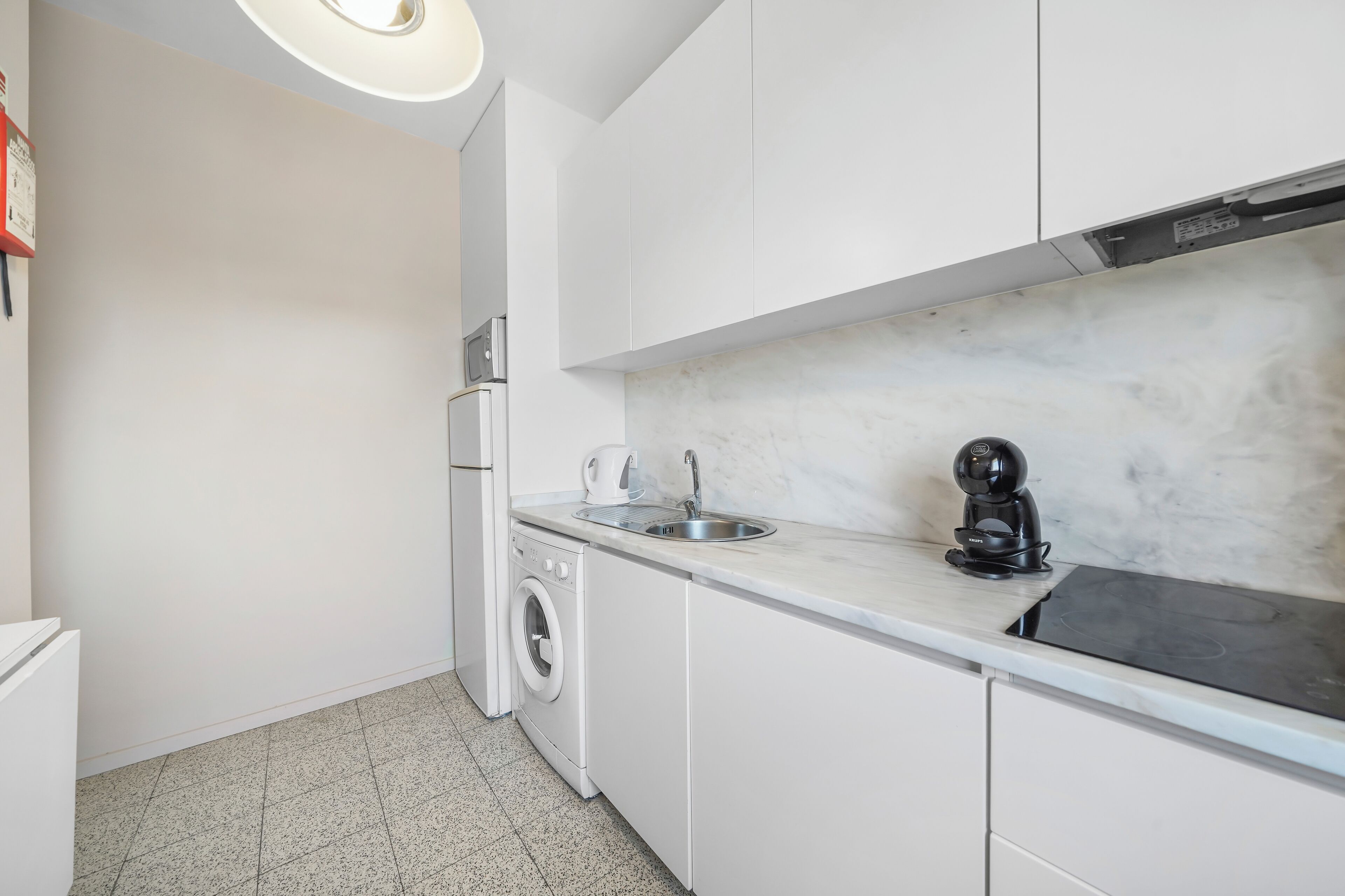 Une cuisine compacte avec des meubles blancs, un lave-linge encastré, un évier et une machine à café.
