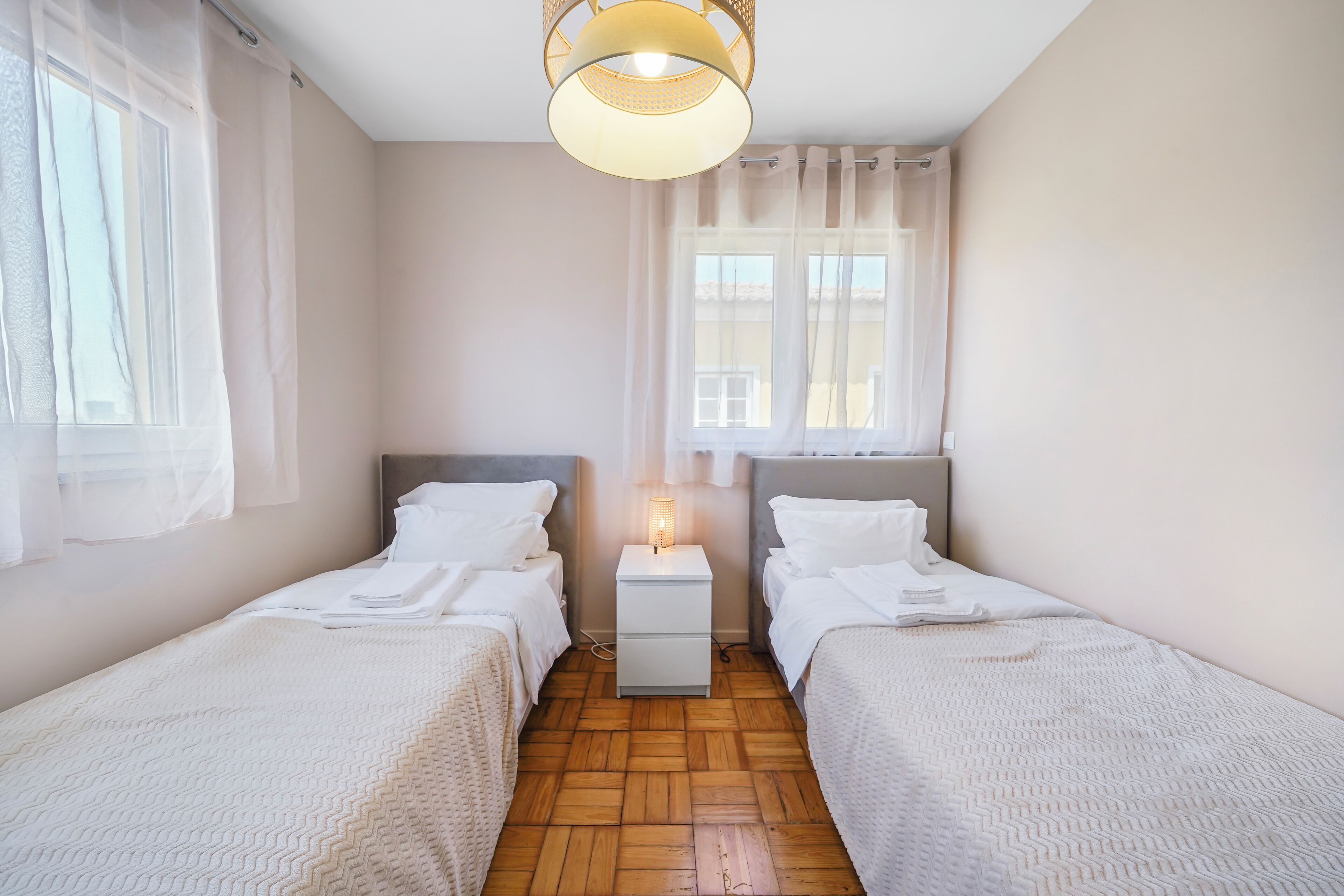 A bedroom with two single beds, a bedside table with a lamp, a window with sheer curtains, and a wooden floor.