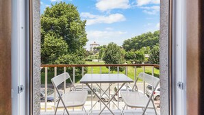 A balcony with two white metal chairs and a table, overlooking a parking area and a green landscape with trees and a building in the distance. - Apartments Luiz I Bridge (Vila Nova de Gaia)