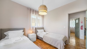 A bedroom with two beds, a bedside table with a lamp, a window with curtains, and a wooden floor. - Apartments Luiz I Bridge (Vila Nova de Gaia)