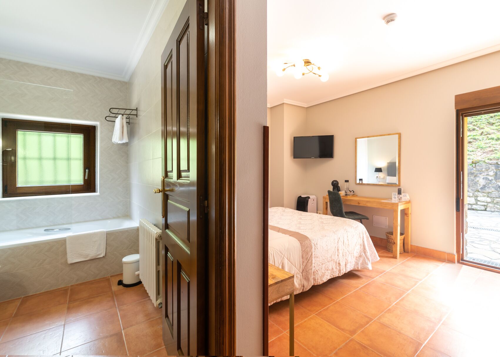 Un baño con bañera, inodoro y ventana, y un dormitorio con cama, escritorio y televisor.