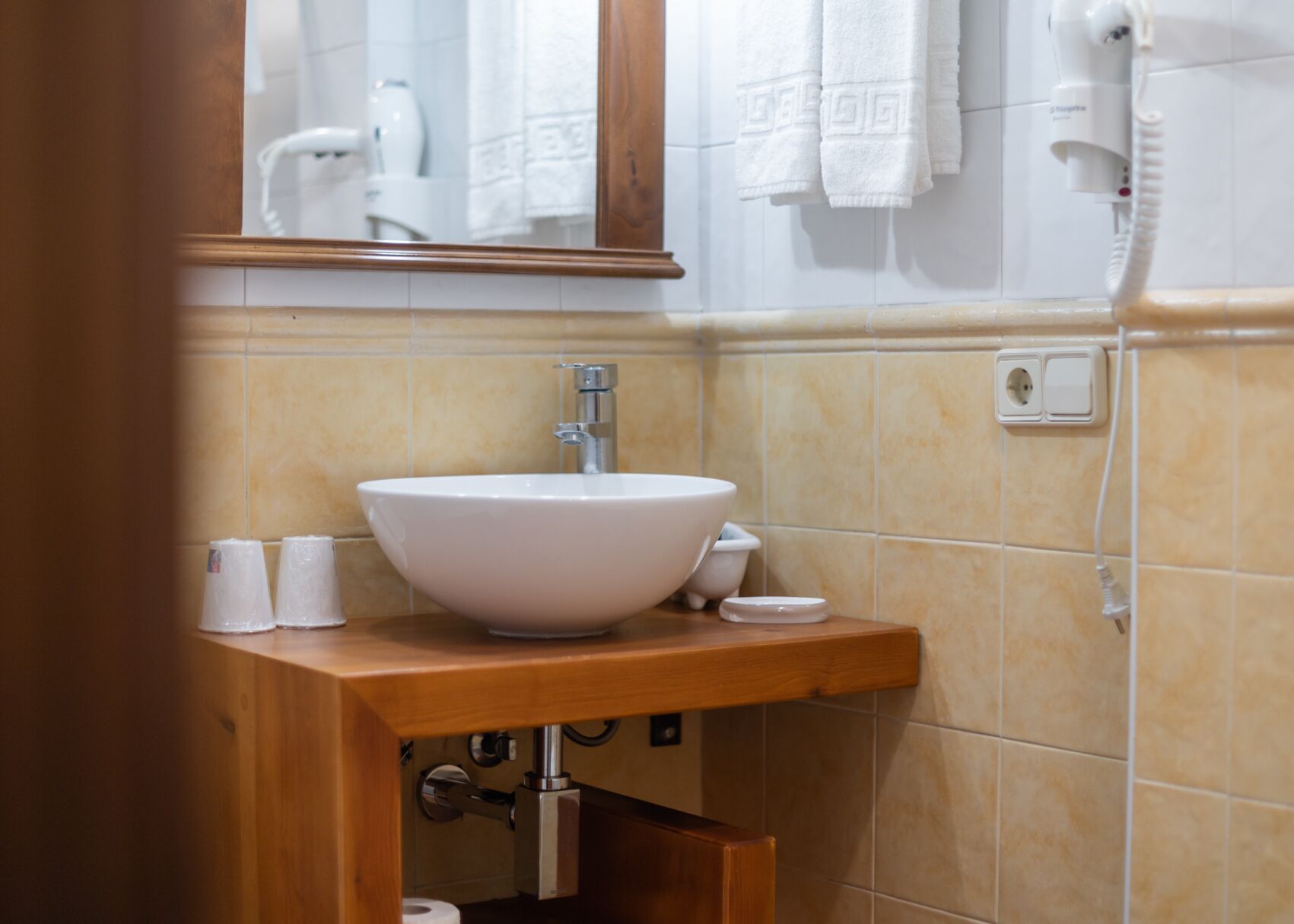 A bathroom with a white sink, wooden vanity, and a mirror above the sink. There are white towels hanging on the wall and a hairdryer on the wall.