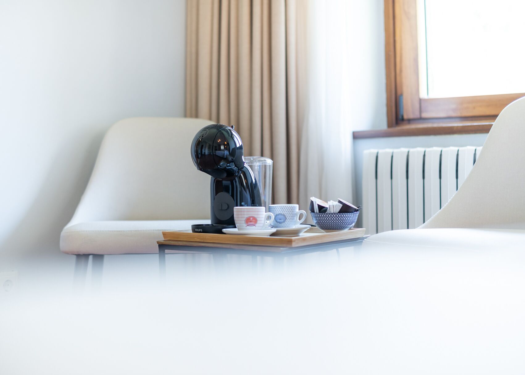 Una habitación de hotel moderna con una elegante cafetera negra sobre una bandeja, un sofá blanco y una ventana con cortinas.