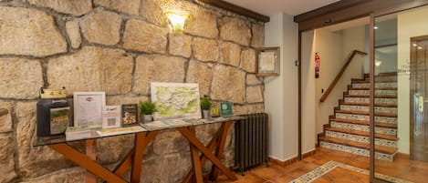 Un muro de piedra, una mesa de madera con folletos, un radiador y una escalera con baldosas.