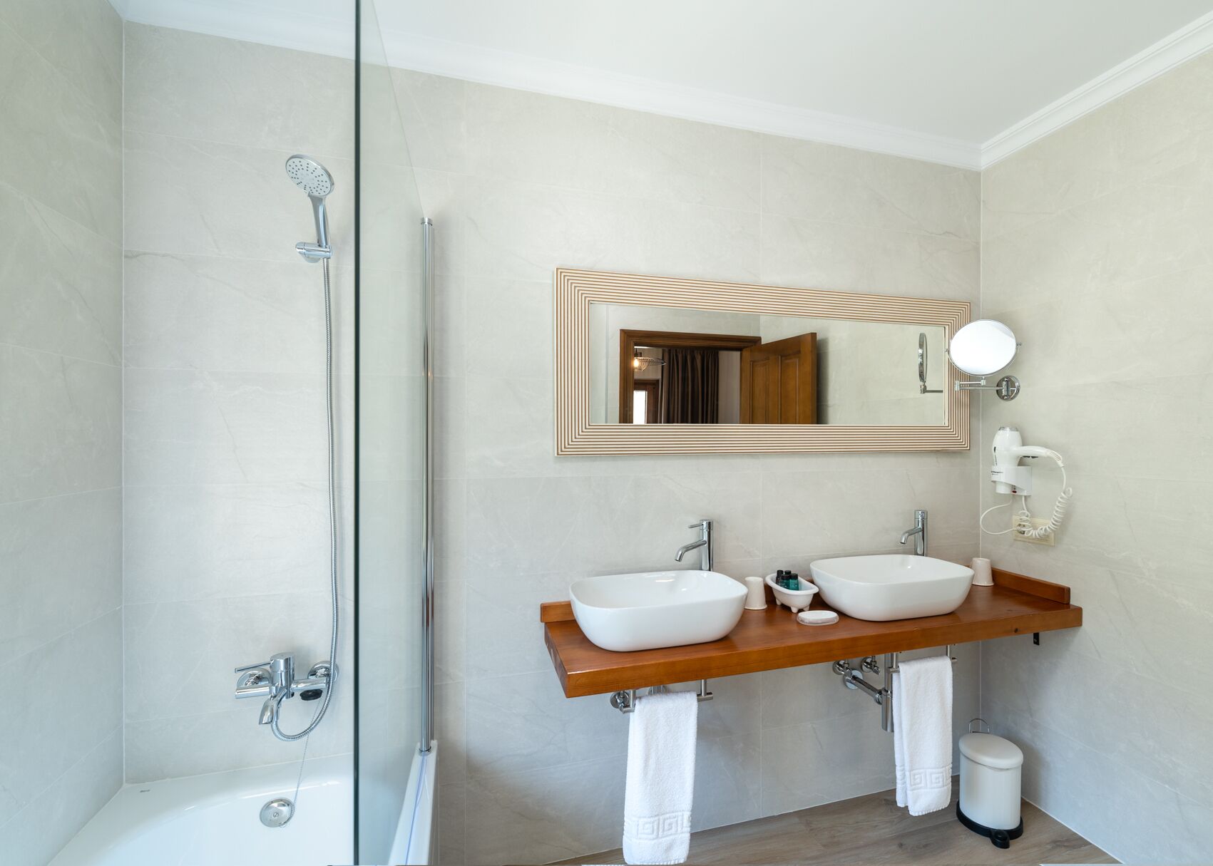 A modern bathroom with a glass shower enclosure, a double sink vanity, a large mirror, and a hairdryer on the wall.