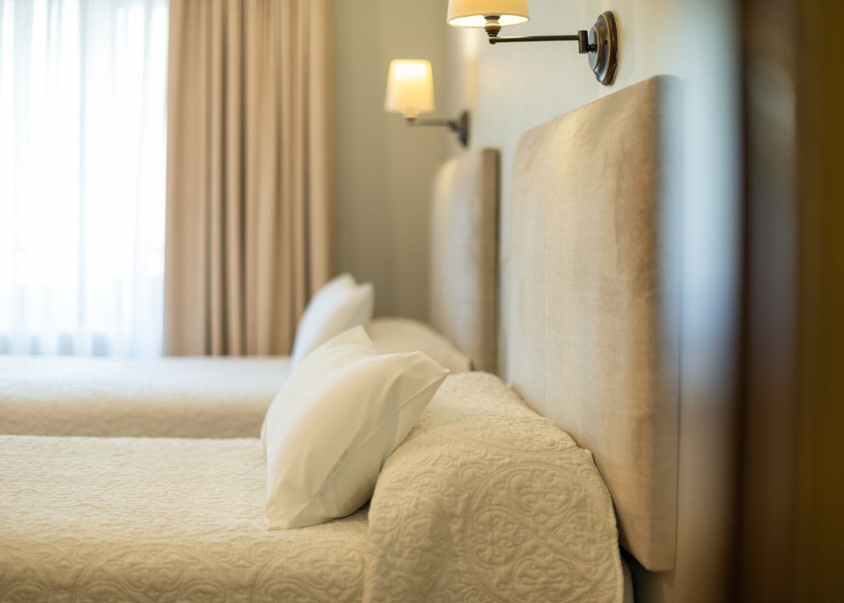 Une chambre d’hôtel avec un lit bien fait, une lampe fixée au mur et une fenêtre avec des rideaux.