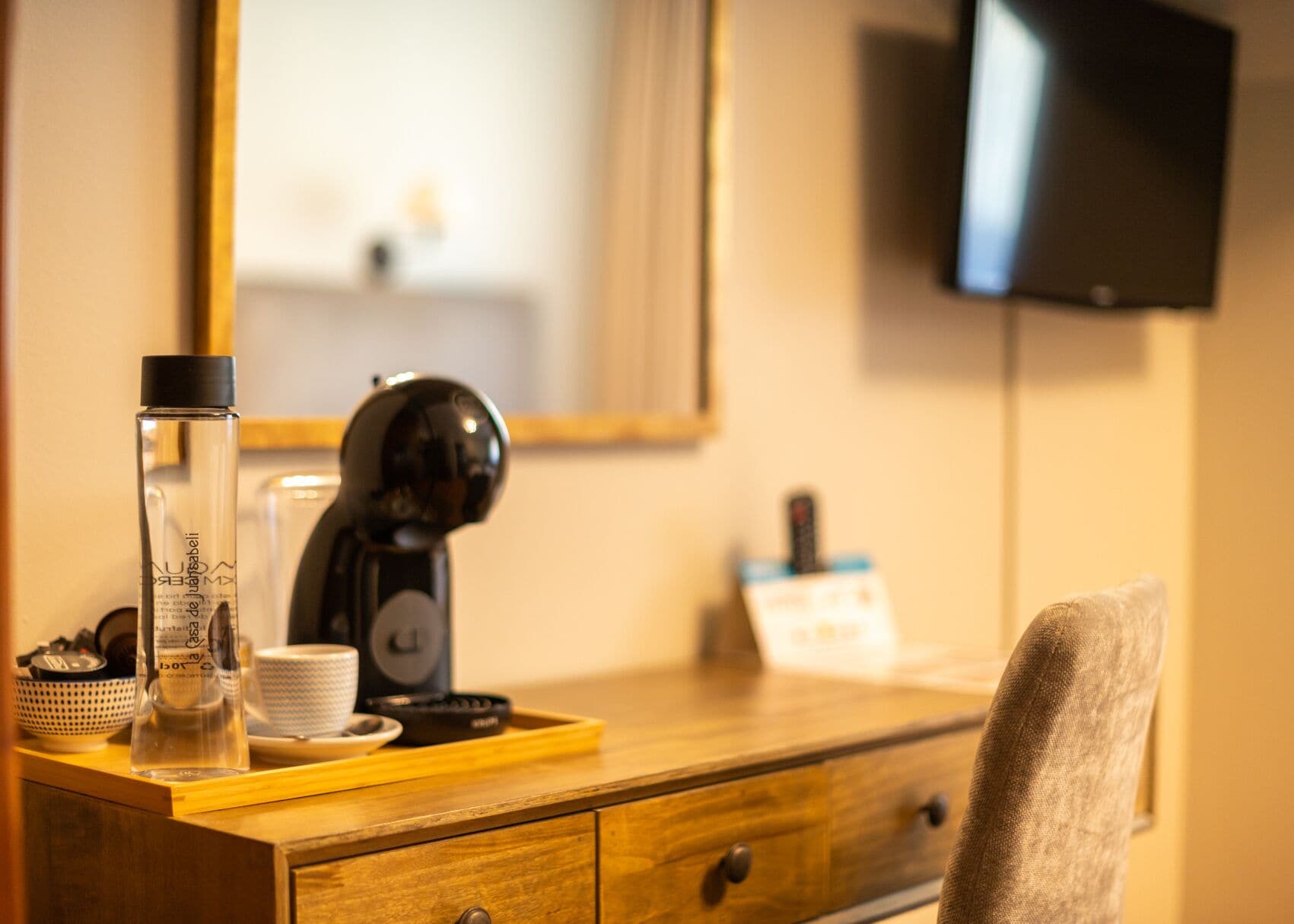 Habitación de hotel con un escritorio de madera, cafetera, botella de vidrio, taza y televisor montado en la pared.