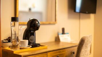 Habitación de hotel con un escritorio de madera, cafetera, botella de vidrio, taza y televisor montado en la pared.