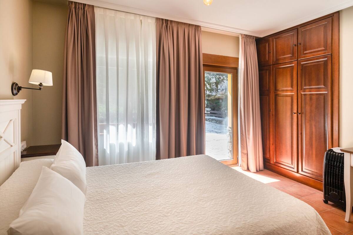 A hotel room with a large bed, a wooden wardrobe, a radiator, and a view of the outdoors.