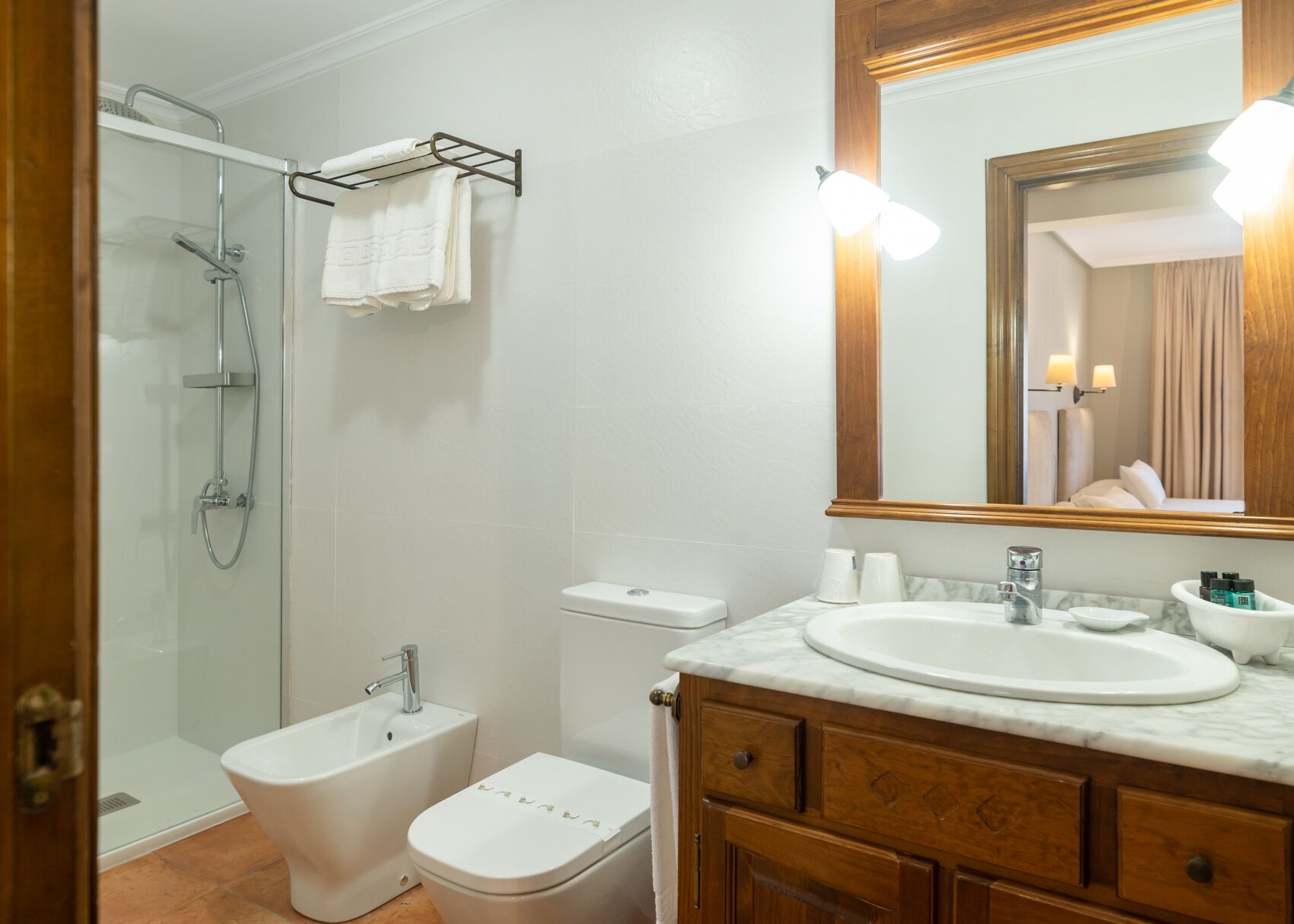 Un baño con ducha de vidrio, inodoro, bidé, lavamanos con encimera de mármol y tocador de madera.