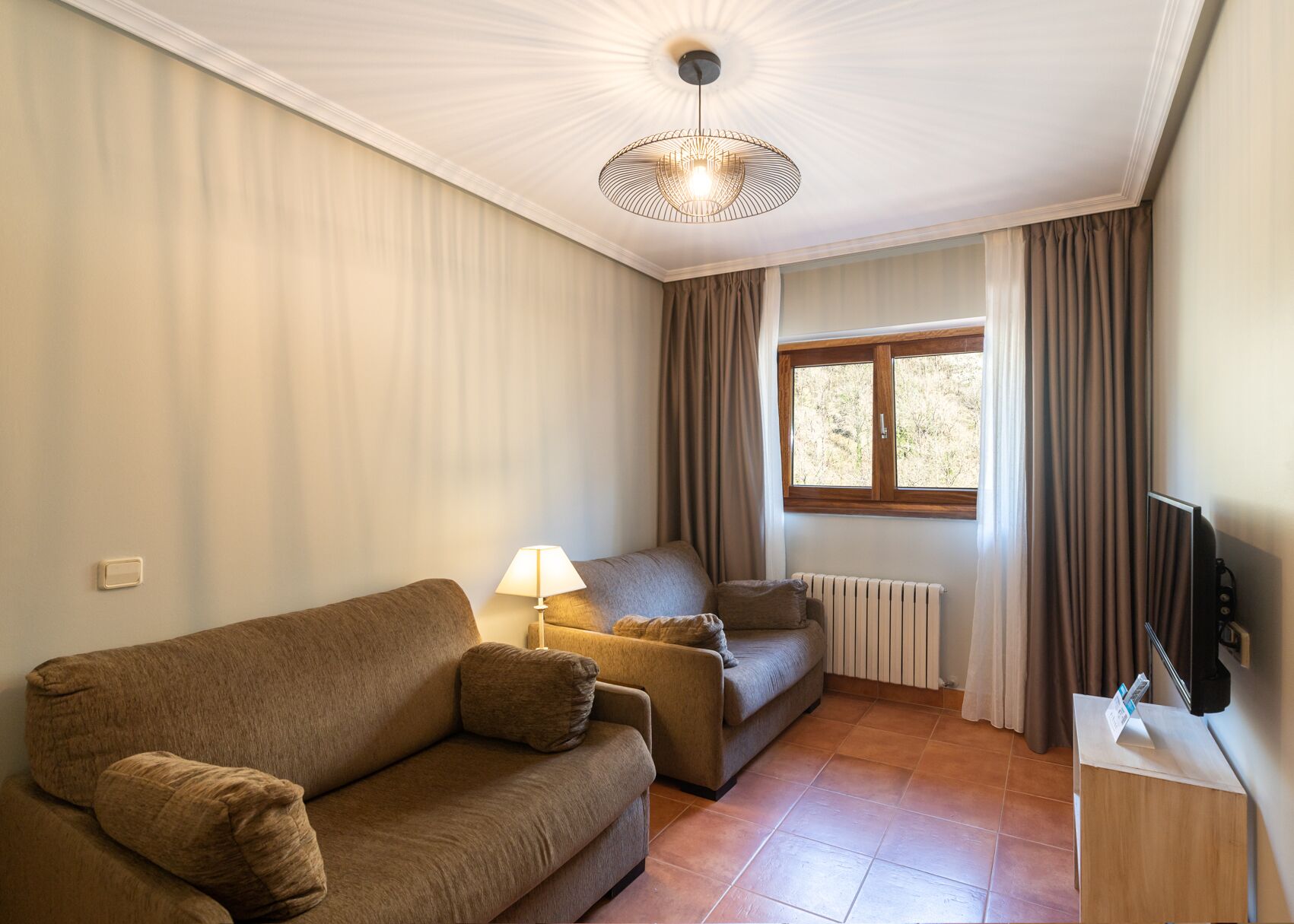 A living room with a sofa, a smaller couch, a lamp, a radiator, a window with curtains, and a television on a stand.