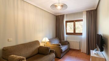 Ein Wohnzimmer mit einer Couch, einem kleinen Sofa, einer Lampe, einer Heizung, einem Fenster mit Vorhängen und einem Fernseher auf einem Schrank.