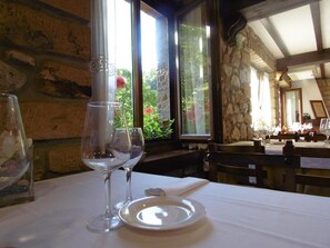 A dining table with a wine glass and a plate, set against a stone wall with a window overlooking a garden. - Logis Hotel Restaurante La Casa de Juansabeli (Cabrales)