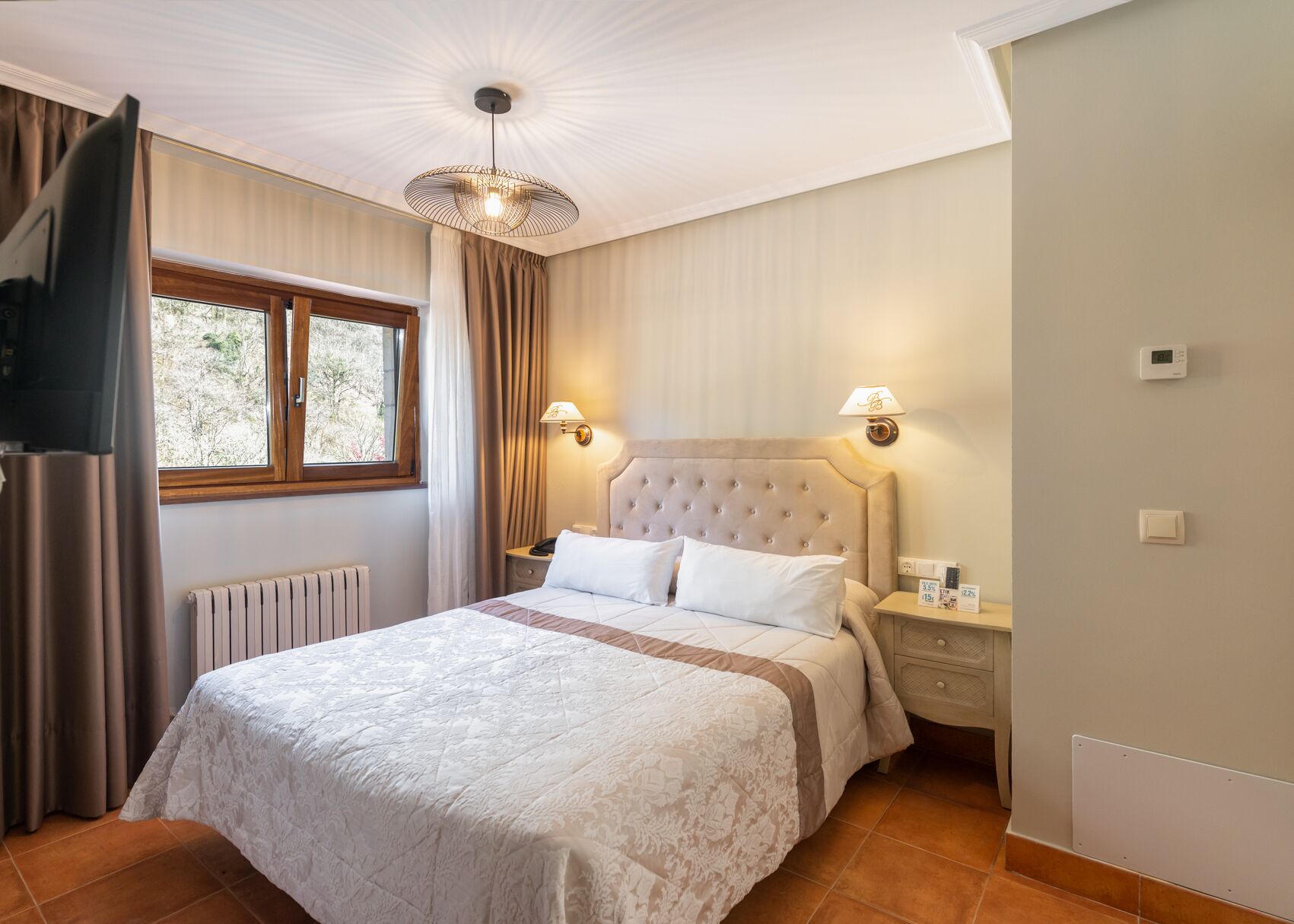 A bedroom with a large bed, a window, a radiator, a wall-mounted light, and a thermostat.