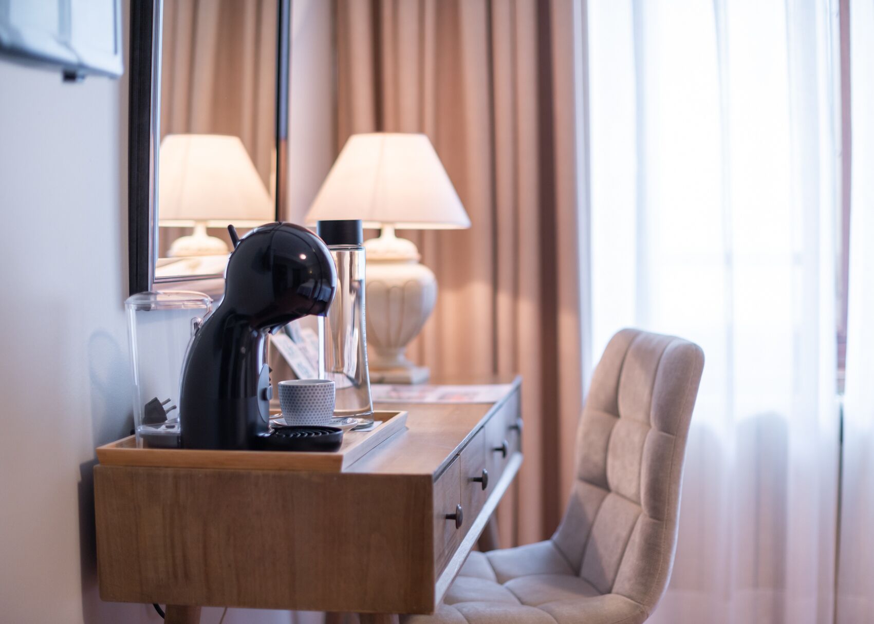 A hotel room with a wooden desk, a black coffee machine, a cup, and a lamp.