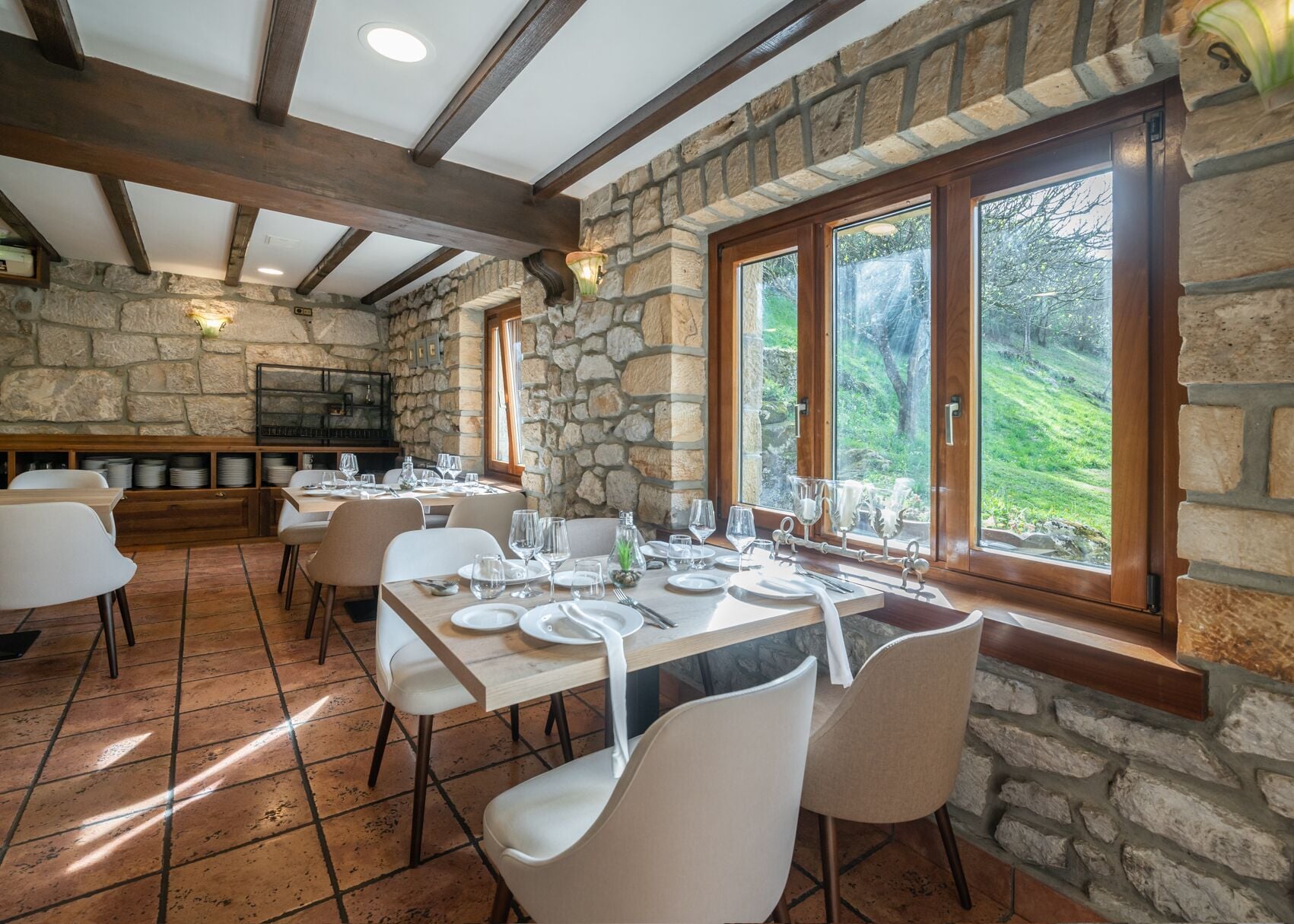 Un restaurante con paredes de piedra, vigas de madera y amplios ventanales que dan a un paisaje verde. Las mesas están dispuestas con manteles blancos y cubiertos.