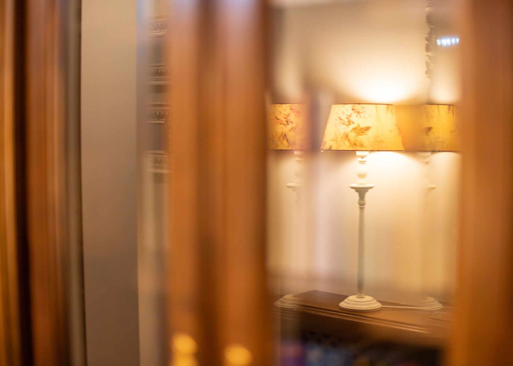 A room with a lamp on a table, curtains partially drawn, and a reflection of a window.