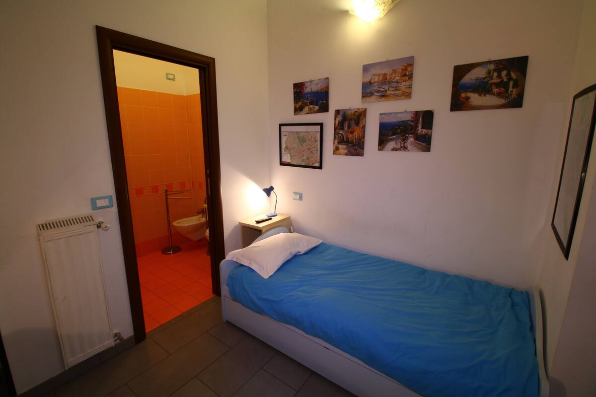 A small bedroom with a single bed, a nightstand, a wall-mounted lamp, and several framed pictures.