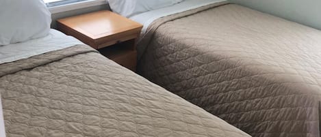 Une chambre avec deux lits, chacun recouvert d’une courtepointe matelassée et agrémenté d’oreillers blancs. Une petite table de chevet en bois se trouve entre les deux lits.