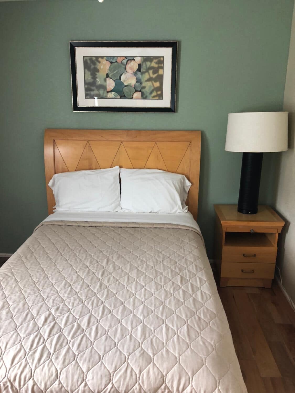 A neatly made bed with a quilted cover, a wooden headboard, a framed artwork above the bed, a bedside lamp, and a wooden nightstand with drawers.