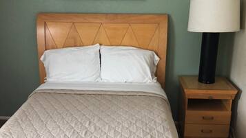 A neatly made bed with a quilted cover, a wooden headboard, a framed artwork above the bed, a bedside lamp, and a wooden nightstand with drawers.