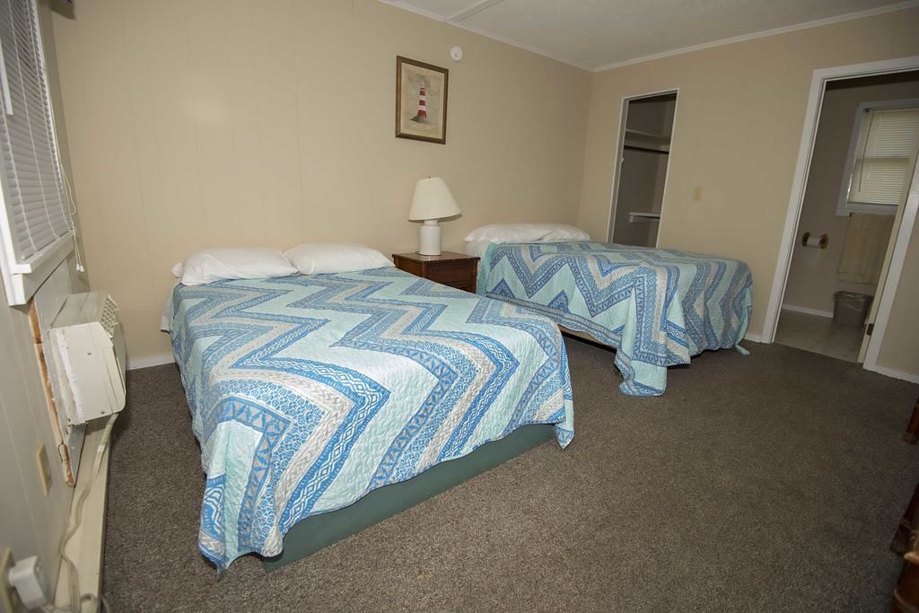 Une chambre d’hôtel avec deux lits, des murs de couleur beige, un tableau encadré, une table de chevet avec une lampe et une porte donnant sur une autre pièce.