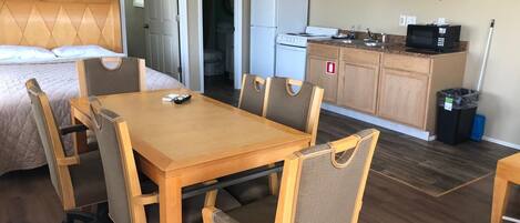 A hotel room with a bed, a wooden dining table with chairs, a kitchenette with a refrigerator, microwave, and coffee maker.