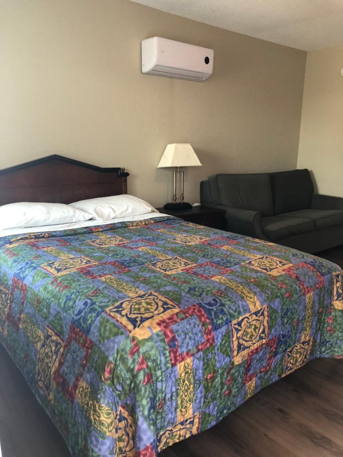 A hotel room with a bed, a couch, a lamp, and an air conditioning unit.