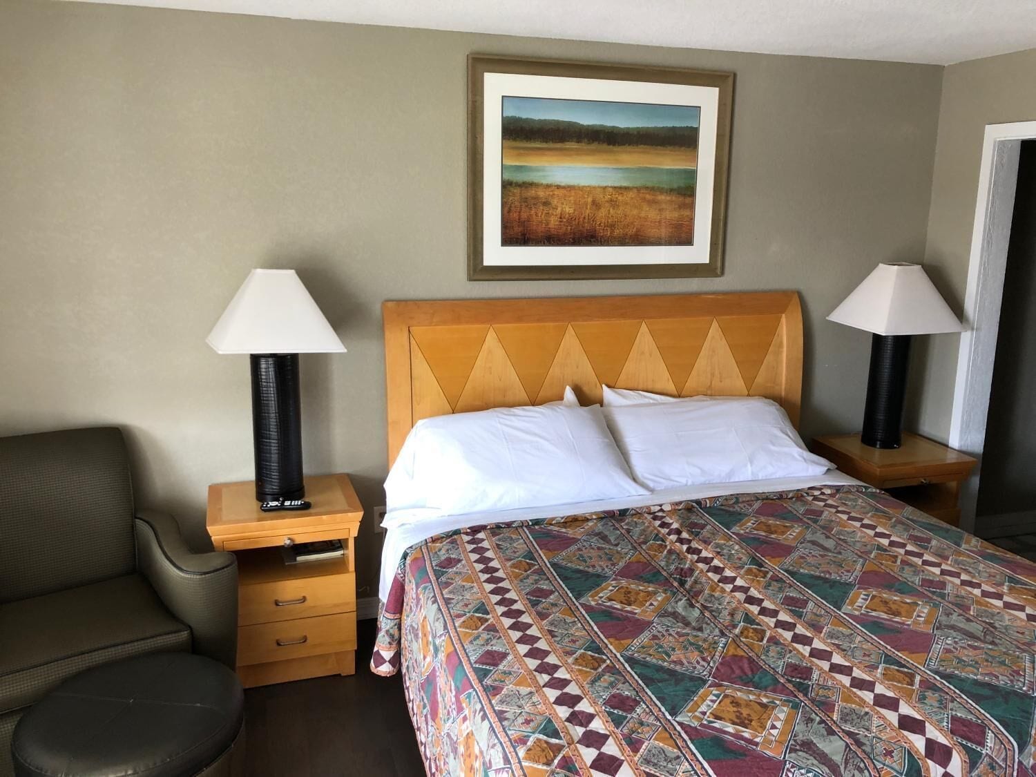 Une chambre d’hôtel avec un lit, deux lampes de chevet, une chaise, une table de chevet en bois et un tableau représentant un paysage, encadré et accroché au mur.
