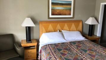 Une chambre d’hôtel avec un lit, deux lampes de chevet, une chaise, une table de chevet en bois et un tableau représentant un paysage, encadré et accroché au mur.