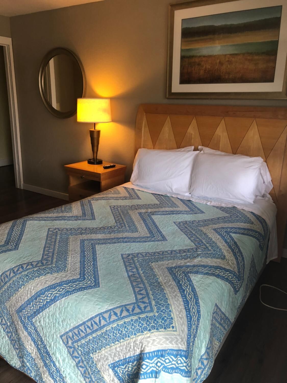 A hotel room with a bed, a nightstand with a lamp, a round mirror, and a painting on the wall.