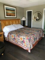 Una cama bien hecha con una colcha estampada, cabecero de madera, lámpara de noche y un cuadro enmarcado en la pared.