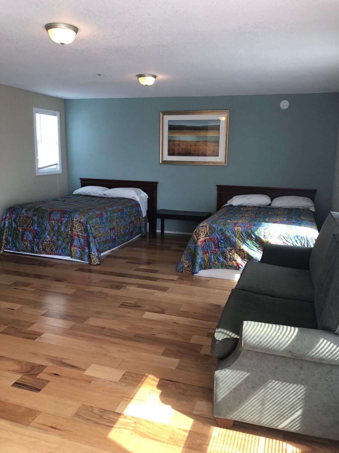 A room with two beds, a sofa, wooden flooring, a painting on the wall, and a window.